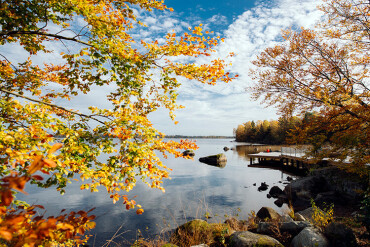 Herbstliche Landschaft mit bunten Laubbäumen an einem ruhigen Seeufer in Stockholm mit Steg und Felsen im Vordergrund
