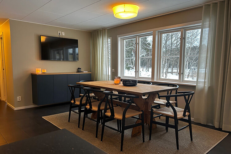 Esszimmer mit Holztisch, sechs schwarzen Stühlen, Teppich, Sideboard mit Fernseher und großen Fenstern mit Blick auf verschneite Bäume