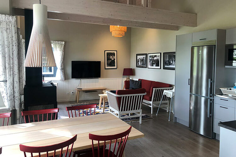Gemütliches Wohnzimmer mit rotem Sofa, weißen Sesseln, großem Fernseher, Esstisch mit roten Stühlen und moderner Küche mit Edelstahlkühlschrank
