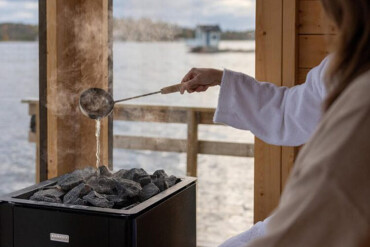 Person in weißem Bademantel gießt Wasser mit einer Kelle auf heiße Steine in einer Sauna mit Blick auf Wasser und Gebäude im Hintergrund