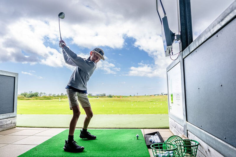 Junge in Golfkleidung beim Abschlag auf einer Golfanlage mit grünem Kunstrasen und offenem Blick auf eine Wiese.