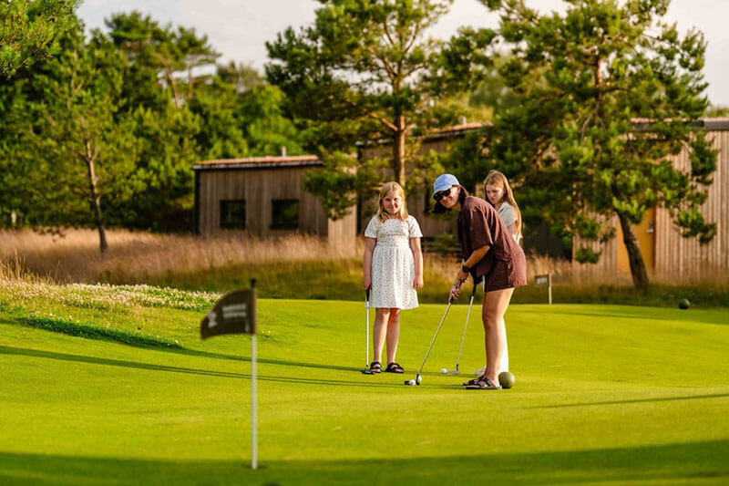 Frau mit Sonnenhut spielt Golfputt auf Grün mit zwei Mädchen im Hintergrund vor Holzhäusern und Kiefern.