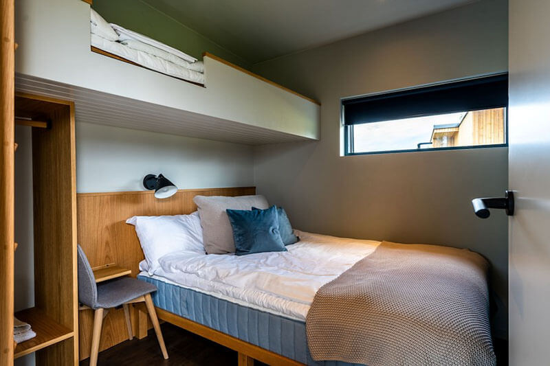 Kleines Schlafzimmer mit Doppelbett, Hochbett, Stuhl, Leselampe und Fenster mit Blick auf ein Holzhaus.