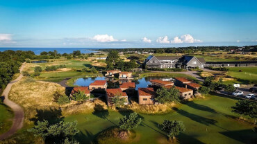 Luftaufnahme eines Golfplatzes mit mehreren kleinen Gebäuden und einem größeren Gebäude im Hintergrund unter blauem Himmel.