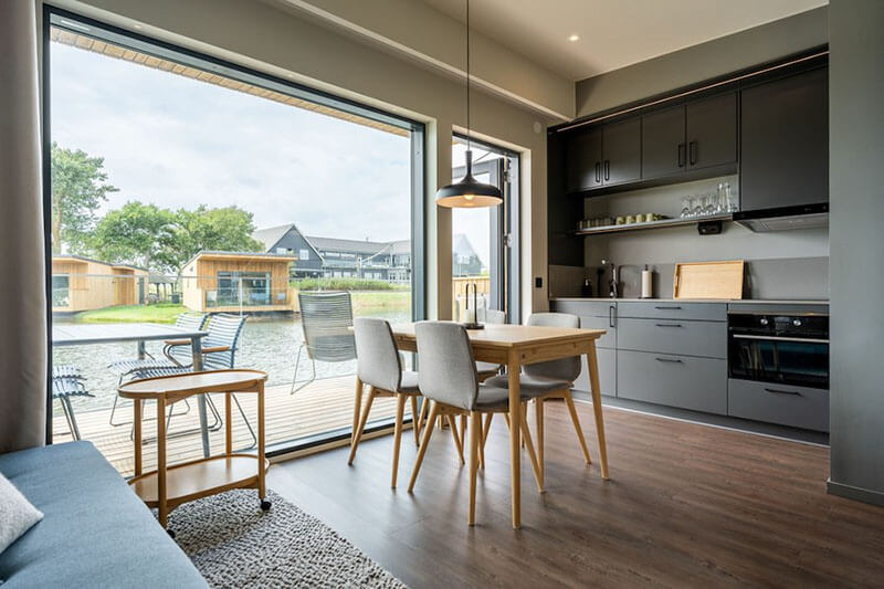 Moderner Essbereich mit Holztisch, Stühlen und Küchenzeile vor großer Fensterfront mit Blick auf Garten und kleine Holzhäuser.