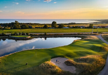 Luftaufnahme eines Golfplatzes mit Wasserhindernis, Sandbunker und Spielern vor einer Küstenlandschaft bei Sonnenuntergang.