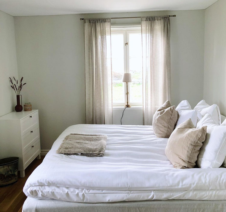 Schlafzimmer mit großem Bett, mehreren Kissen, weißen Vorhängen und Kommode mit Vase.