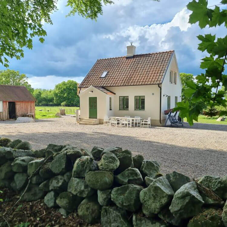 Weißes Ferienhaus mit rotem Ziegeldach hinter einer niedrigen Steinmauer in ländlicher Umgebung unter bewölktem Himmel.