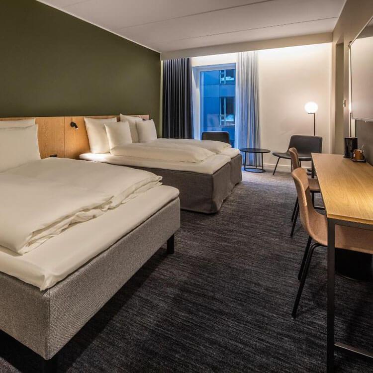 Hotelzimmer mit zwei großen Betten, Schreibtisch mit Stuhl, Sitzbereich mit zwei Sesseln und kleinem Tisch vor einem Fenster.