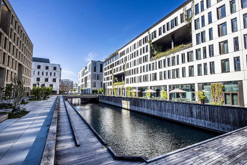 Moderner Gebäudekomplex mit rechteckigen Fenstern entlang eines schmalen Kanals und Holzsteg im Vordergrund.
