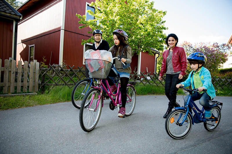 Familie mit zwei Kindern fährt auf Fahrrädern auf einer Straße neben einem roten Haus und einem Holzzaun.