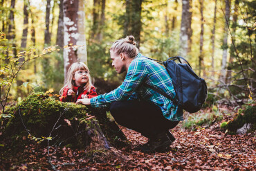 Vater mit Rucksack hockt im Wald vor kleinem Kind auf Baumstumpf.