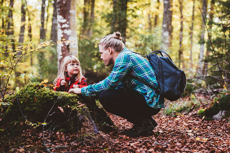 Vater mit Rucksack hockt im Wald vor kleinem Kind auf Baumstumpf.