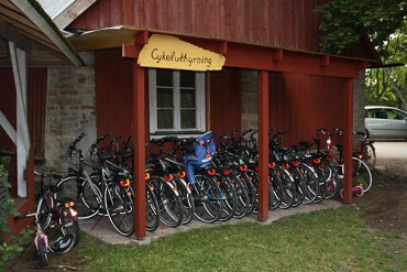 Überdachter Fahrradständer mit mehreren schwarzen Fahrrädern und einem Kindersitz vor einem roten Holzgebäude mit dem Schild 'Cykeluthyrning'