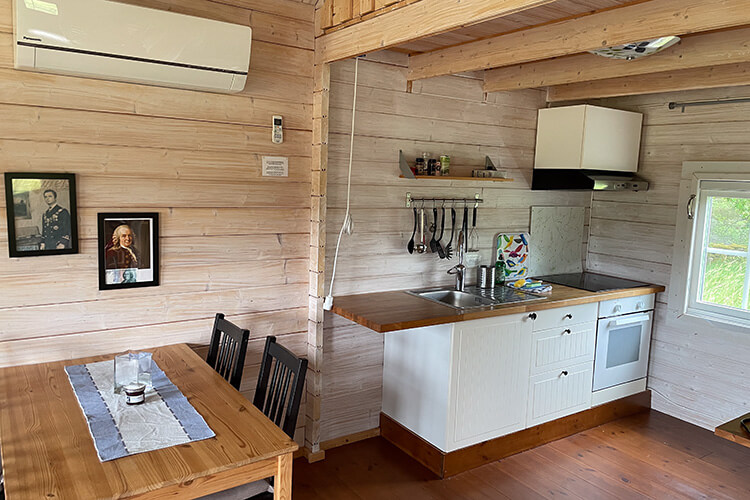 Kleine Landhausküche mit Holztisch und Stühlen, Holzverkleidung an den Wänden, zwei gerahmte Porträtbilder an der Wand und Küchenzeile mit Spüle, Herd und Dunstabzug.
