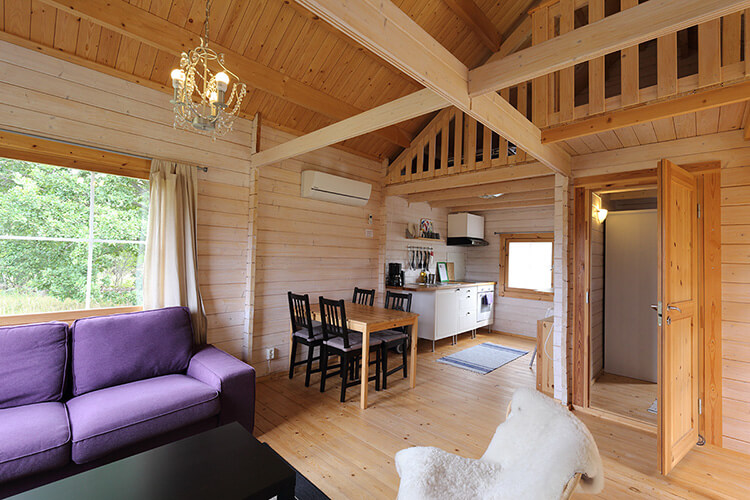Holzferienhaus mit sichtbaren Holzbalken, lila Sofa, Esstisch mit vier Stühlen und offener Küche in einem Raum
