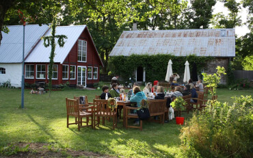 Gruppe von Menschen sitzt an einem langen Holztisch im Garten eines Bauernhofs mit roten und weißen Gebäuden im Hintergrund