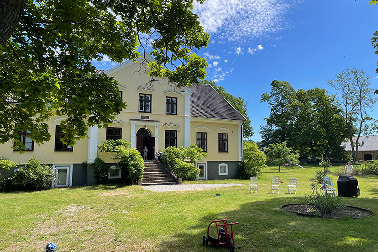Großes gelbes Gutshaus mit Treppe zum Eingang, umgeben von Rasen, Bäumen und Gartenmöbeln bei sonnigem Himmel