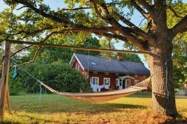 Hängematte zwischen Baum und Holzgestell vor einem roten schwedischen Bauernhaus mit weißen Fensterrahmen und Wäscheleine im Garten