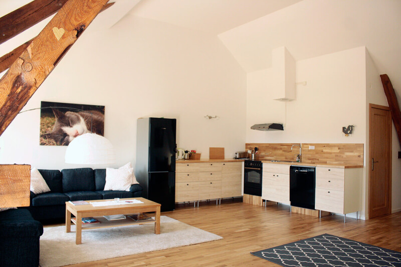 Offene Wohnküche mit Holzboden, weißen Schränken, schwarzem Kühlschrank, schwarzem Sofa, Holztisch und Dachbalken aus Holz in einem hellen Raum in Schweden.