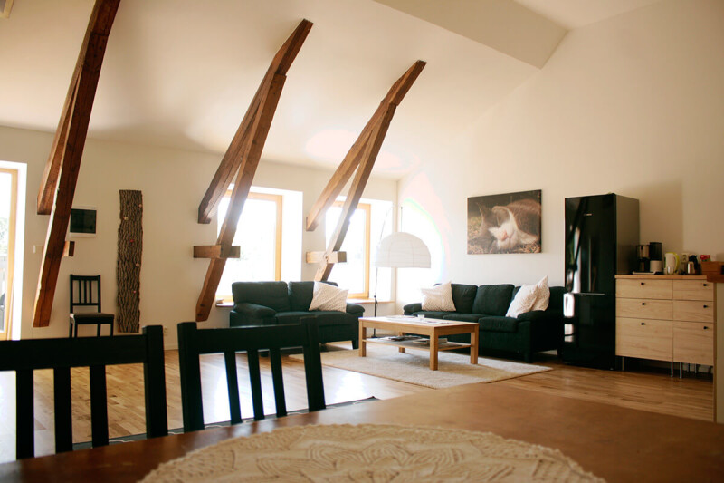 Wohnzimmer mit sichtbaren Holzbalken, zwei Sofas, einem Couchtisch, einem Teppich, einem großen Stehlampen und einer Kommode in einem hellen Raum mit Fenstern