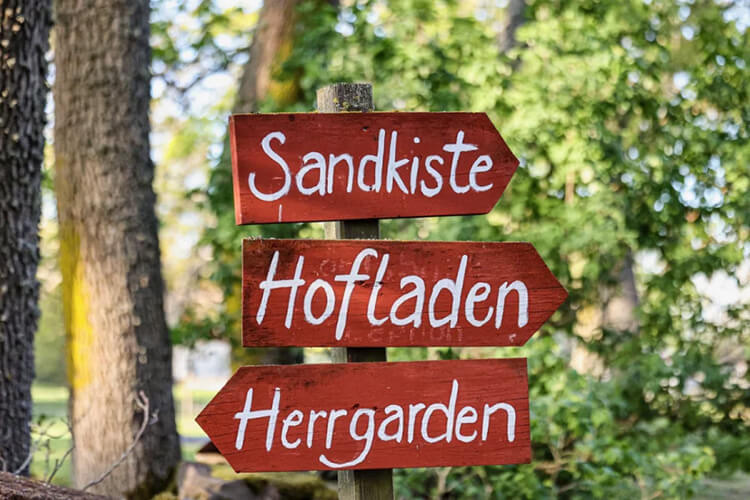Holzwegweiser mit drei roten Schildern und weißer Schrift mit den Aufschriften 'Sandkiste', 'Hofladen' und 'Herrgarden' vor Bäumen im Hintergrund