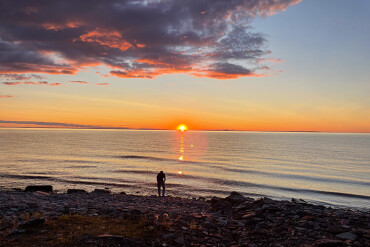 Person steht am felsigen Strand und blickt auf die untergehende Sonne über dem ruhigen Meer in Schweden