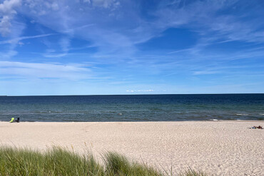 Strand mit hellem Sand, im Vordergrund Dünen mit Gras, weiter hinten ruhiges Meer unter blauem Himmel mit wenigen Wolken