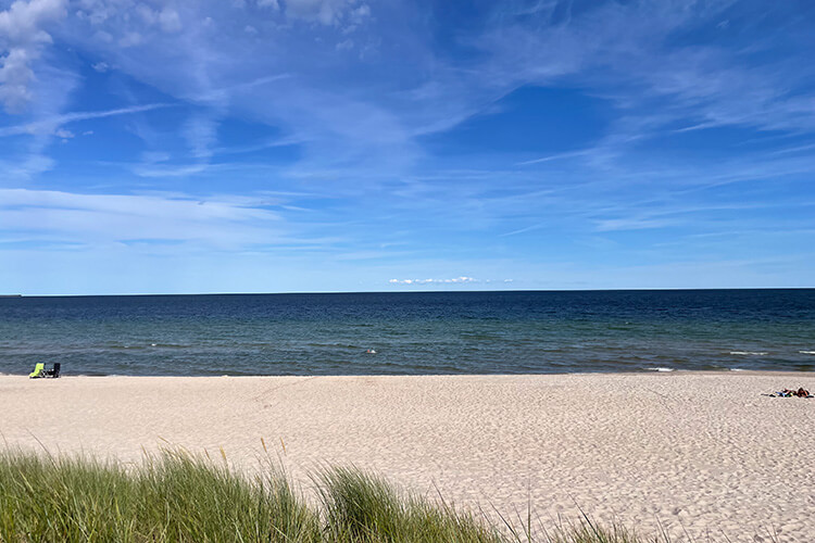 Strand mit hellem Sand, im Vordergrund Dünen mit Gras, weiter hinten ruhiges Meer unter blauem Himmel mit wenigen Wolken