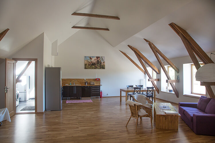 Großer offener Wohnraum mit sichtbaren Holzbalken, Küchenzeile, Esstisch, Sofa und Stühlen in einem hellen Dachgeschoss einer Wohnung auf einem Familienbauernhof in Schweden