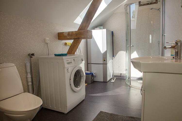 Badezimmer mit Toilette, Waschmaschine, Waschbecken, Dusche und sichtbarem Holzbalken unter einer Dachschräge in einer Wohnung auf Öland, Schweden