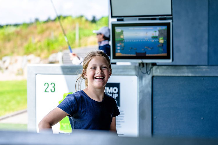 Lachendes Mädchen in blauem T-Shirt steht an einer Golf-Driving-Range mit Monitor im Hintergrund.
