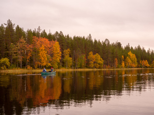 Ein Kanu fährt am Ufer eines Sees entlang. Am Ufer stehen Bäume in herbstlichen Farben.