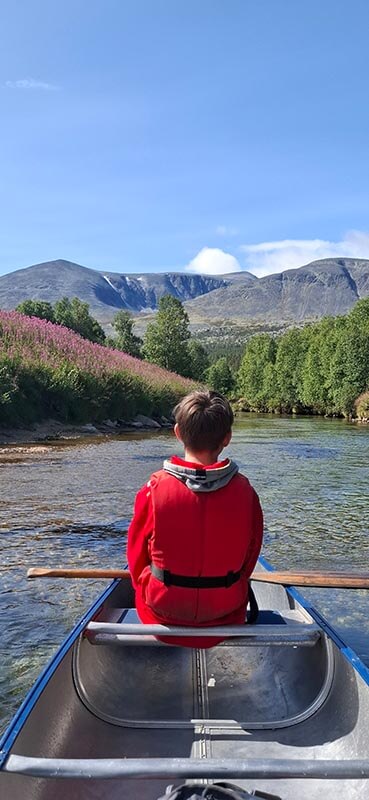 Kind mit rotem Oberteil sitzt in einem Kanu auf einem Fluss, umgeben von grünen Bäumen und Bergen im Hintergrund