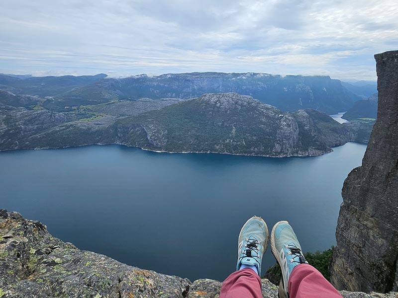 Blick von einem Felsen auf einen tiefblauen Fjord mit umliegenden Bergen und zwei ausgestreckten Beinen mit Sportschuhen im Vordergrund