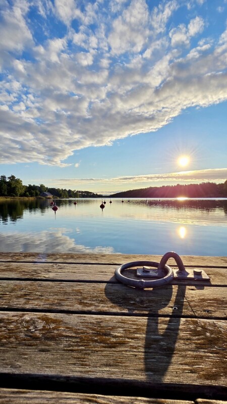 Ein Sommerliches Bild von einem Holzsteg aus fotografiert mit Wolken und Sonne, die sich im Wasser spiegeln.